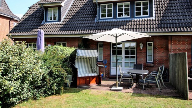Une maison avec un toit en tuiles, une façade en briques, un patio couvert avec une table et des chaises, et un grand parasol.