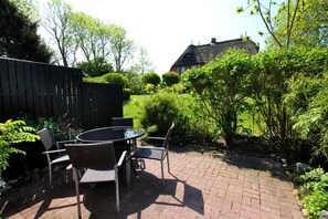 En terrasse med et rundt bord og stole, omgivet af en murstenssti og frodig vegetation.
