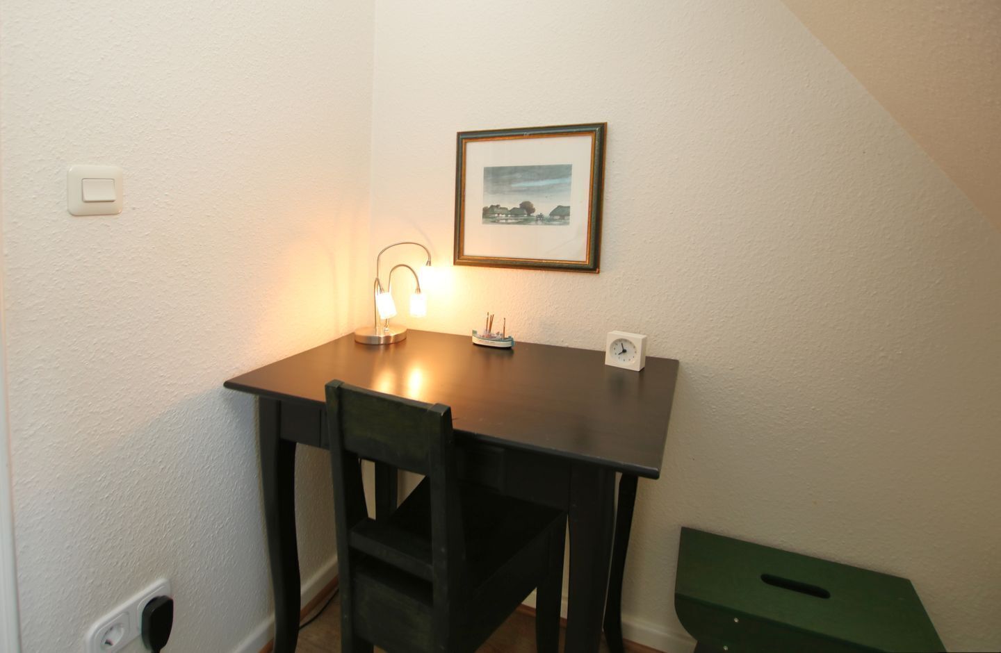 A small, dark wooden table with two chairs, a framed picture on the wall, a desk lamp, and a clock.