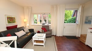 A living room with a black leather sofa, a white coffee table, a beige armchair, and a white cabinet. There are two windows with curtains, a door, and a framed picture on the wall. - Ohl Dörp 22 Pastoratshof, Haus 3 - Ferienwohnung in Wrixum, mit 3- Zimmern auf 60m² im Reetdachhaus (Wrixum)