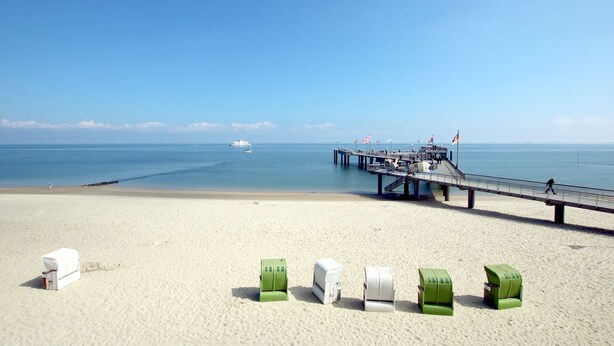 A beach with a pier extending into the water, featuring a small boat and flags. - Sandwall 11a, Whg. 3 "gästehaus Gorch Fock" - Ferienwohnung mit 2- Zimmern auf ca. 42m² mit Direktem (Wyk auf Föhr)