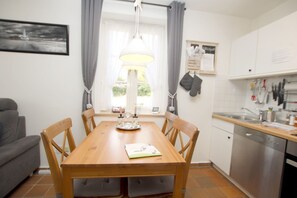 A dining area with a wooden table, four chairs, a pendant light, a window with curtains, a kitchen area with a sink and utensils, and a framed picture on the wall.