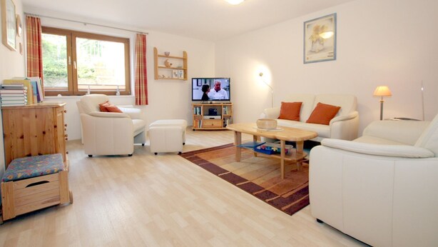 Een woonkamer met een bank, een voetenbankje, salontafel, televisie, boekenplank en een raam met gordijnen.