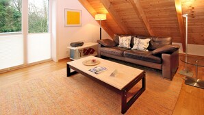 Une chambre à coucher confortable, aménagée en style « loft », avec un plafond en bois, un canapé en cuir marron, une petite table d’appoint et une grande fenêtre.