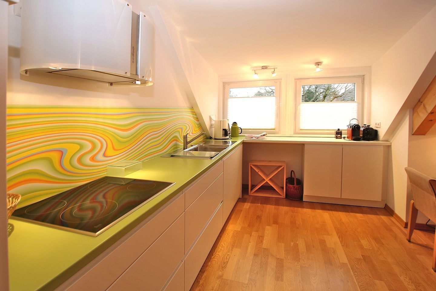 Una cocina con un salpicadero colorido, una estufa y un lavabo. Hay dos ventanas con persianas, un cesto de madera y una silla.