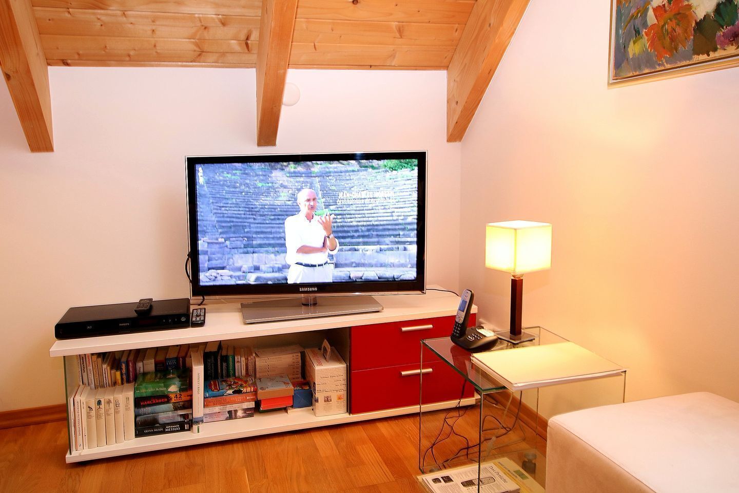 Ein Wohnzimmer mit einem Flachbildfernseher auf einem Holzständer, einem Bücherregal mit Büchern, einem roten Schrank, einem Glastisch mit einer Lampe und einer Holzzahl.