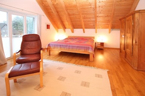 Quarto com teto de madeira, cama com manta colorida, poltrona de couro e guarda-roupa de madeira.