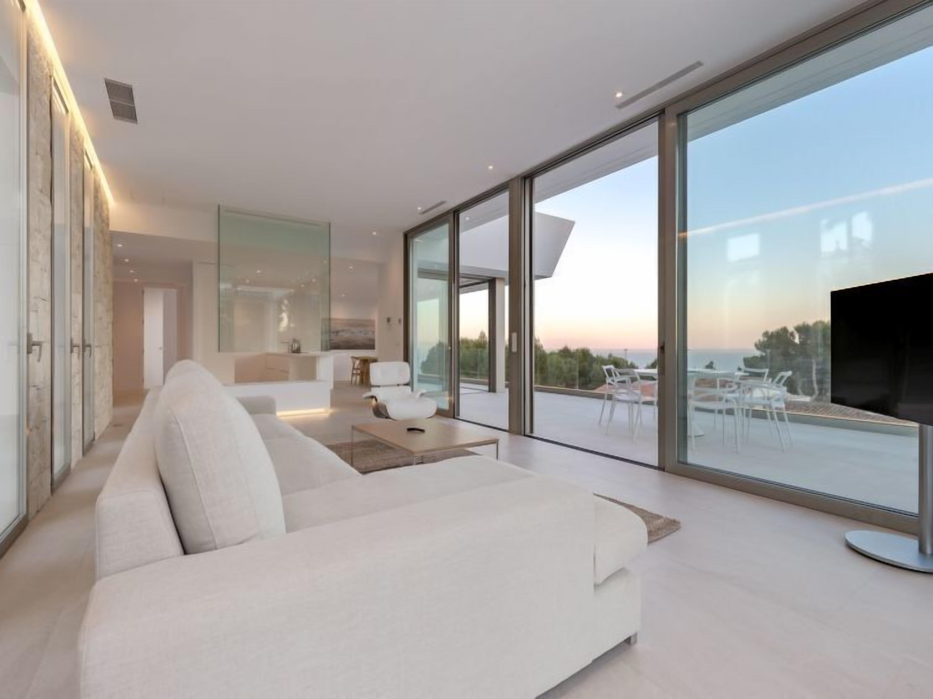 A modern living room with a large sofa, a flat-screen TV, and a balcony with a view of the sea.