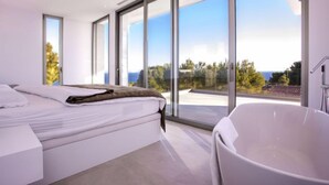 Ein modernes Schlafzimmer mit einem großen Bett, einer Badewanne und raumhohen Fenstern mit Blick ins Freie.