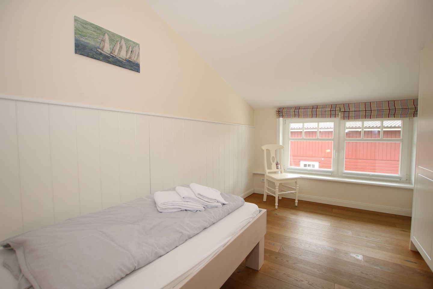 A single bed with a white headboard, a chair, a window with a plaid valance, and a painting on the wall.