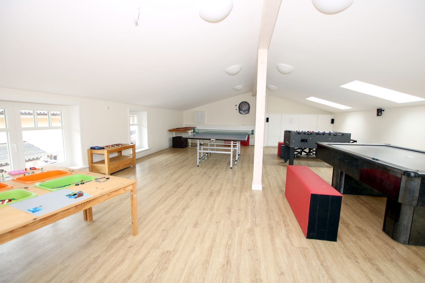 A spacious room with a pool table, foosball table, and a ping pong table. There are wooden tables with toys and a window providing natural light.