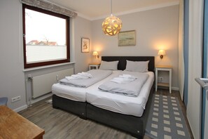 A double bed with white linens, two bedside tables with lamps, a wooden floor, and a window with blinds. - Gmelinstraße 5 a, Whg. A3, By de Wyk - Holiday apartment. Wyk, 2 rooms, approx. 52 m² (Wyk auf Föhr)