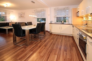 A modern kitchen with wooden flooring, a dining area with a glass table and black chairs, and a view of the outside through a window. - Hasenkamp 1 - Ferienwohnung mit 4- Zimmern auf ca. 75m² (Wyk auf Föhr)