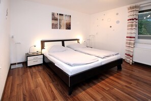 A neatly made bed in a room with a wooden floor, a nightstand, a wall clock, and a window with curtains. - Hasenkamp 1 - Ferienwohnung mit 4- Zimmern auf ca. 75m² (Wyk auf Föhr)