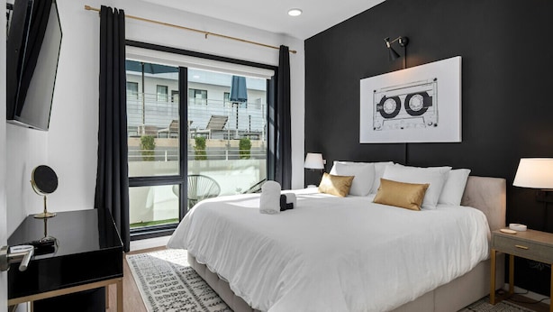 A modern hotel room with a large bed, a flat-screen TV, a desk with a mirror, a framed picture on the wall, and a view of the outside. - UPSCALE Cozy Nashville Condo★ POOL, PATIO, PARKING (Nashville)
