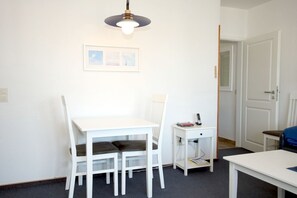 Ein Zimmer mit einem weißen Tisch, zwei Stühlen, einem kleinen weißen Beistelltisch mit Telefon und Buch sowie einer Deckenleuchte.