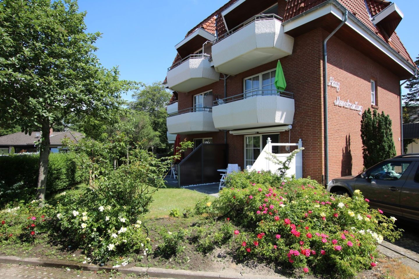 A multi-story building with balconies, a garden with flowers, and a parked car.