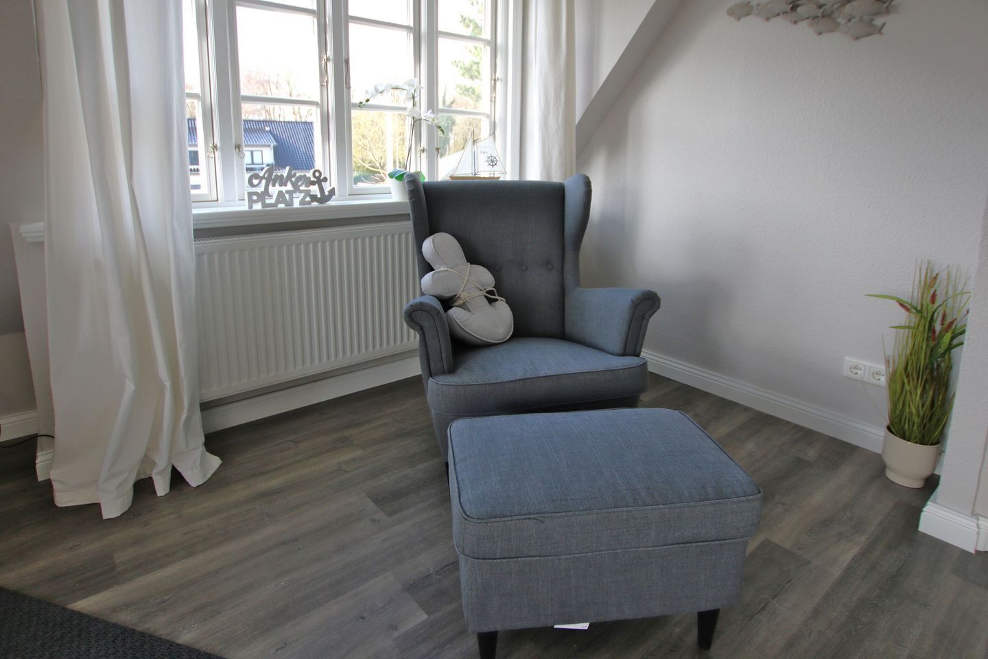 Una habitación con un sillón tapizado gris y un reposapiés, un radiador, una ventana con cortinas blancas y una planta en maceta.