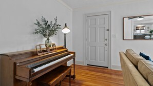 A living room with a wooden piano, a lamp, a plant, and a beige sofa.