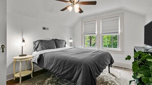 A bedroom with a bed, a ceiling fan, a bedside table with a lamp, a window with blinds, and a television on a stand.