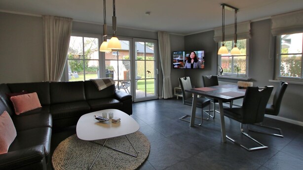 Ein modernes Wohnzimmer mit einem schwarzen Ledersofa, einem Couchtisch und einem Esstisch mit Stühlen. Ein Fernseher ist an der Wand montiert und große Fenster bieten einen Blick ins Freie.