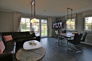A modern living room with a black leather sofa, a coffee table, and a dining table with chairs. There is a TV mounted on the wall and large windows providing a view of the outdoors.