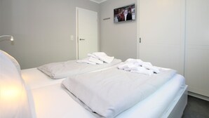 A hotel room with two beds, white linens, a flat-screen TV on the wall, and a door with a handle. - Wiesenweg 4, Whg. 1, Wiesenkieker - Ferienwohnung mit 3- Zimmern auf ca. 76m² (Wyk auf Föhr)