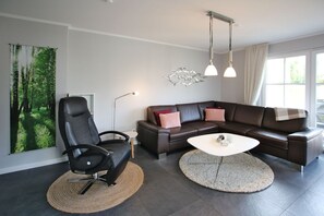 Un salon moderne avec un canapé en cuir, un fauteuil noir, une table basse ronde et une grande œuvre d’art murale sur le thème de la forêt.