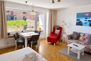 Moderni hotellihuone, jossa on ruokapöytä, tuoleja, punainen nojatuoli, sohva, sänky ja taulu, jossa on kuva auringonlaskusta meren yllä.