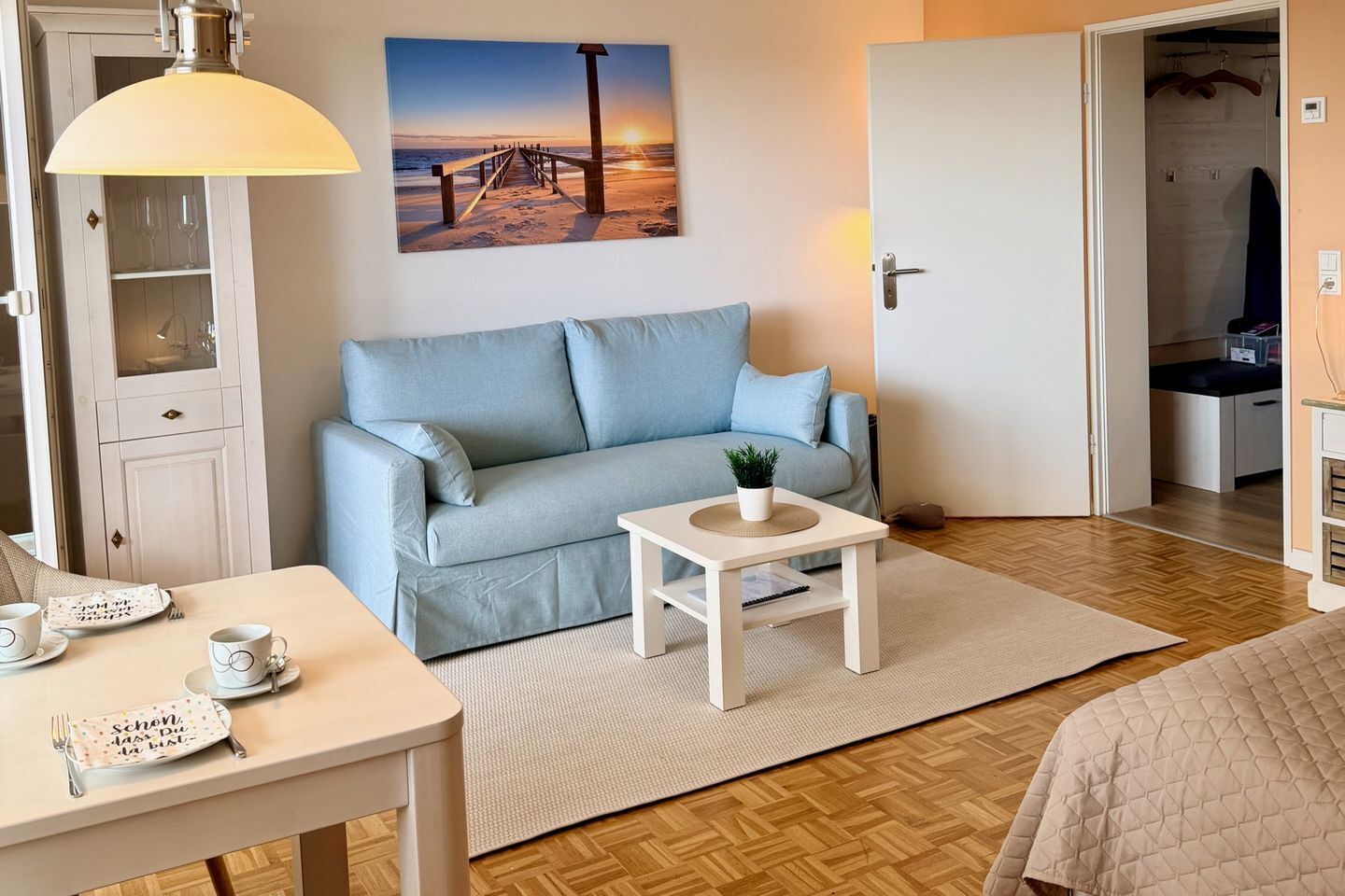 Ein Zimmer mit einer Couch, einem kleinen Tisch mit einer Pflanze, einem Esstisch mit Tassen und einer Tischmatte sowie einem Bild einer Strandansicht.