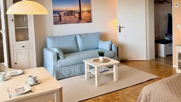 Ein Zimmer mit einer Couch, einem kleinen Tisch mit einer Pflanze, einem Esstisch mit Tassen und einer Tischmatte sowie einem Bild einer Strandansicht.