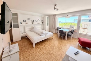 A bedroom with a bed, bedside tables, a desk with chairs, a TV, and a view of the outside.