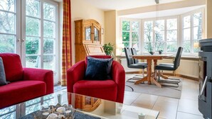 A room with two red armchairs, a glass coffee table, a wooden cabinet, a dining table with chairs, and large windows with a view of trees. - Strandstraße 77a, Haus Nordseeluft - House half with 120m² over 3 floors (Wyk auf Föhr)
