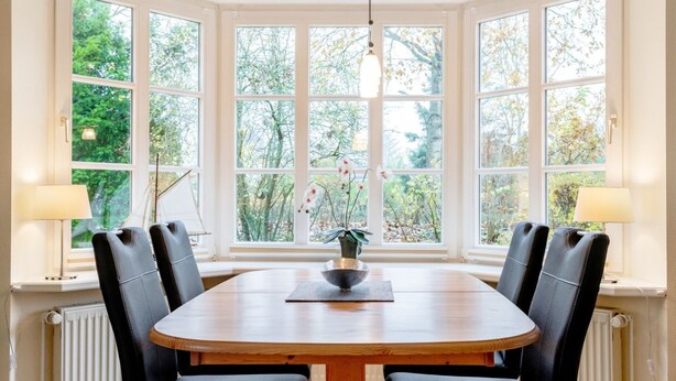 A round wooden table with black leather chairs, a central vase with flowers, and a view of trees through large windows. - Strandstraße 77a, Haus Nordseeluft - House half with 120m² over 3 floors (Wyk auf Föhr)