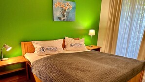 Une chambre d’hôtel avec un mur vert, un tableau représentant des fleurs, deux oreillers ornés du logo BAECKER, une lampe de chevet et un lit recouvert d’une courtepointe matelassée.