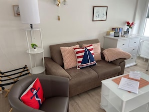 Un salon avec un canapé marron, une table basse blanche, un coussin rayé, un coussin rouge et une petite plante posée sur une table d’appoint blanche.