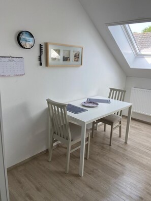 Un comedor con una mesa blanca, dos sillas, un cuadro enmarcado, un reloj de pared y un calendario.