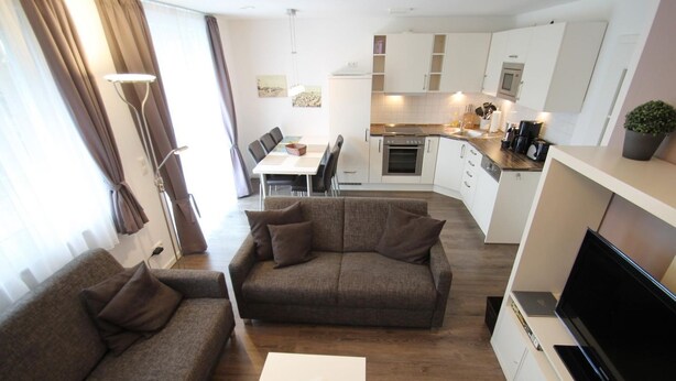 A modern living room with a sofa and a TV, a dining area with a table and chairs, and a kitchen with white cabinets and appliances. - 1012 - Holiday apartment Strandburg - 1012 Holiday apartment Strandburg (Dahme)
