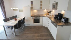 A compact kitchen with white cabinets, a built-in oven, a microwave, and a coffee maker. There is a dining table with chairs and a small rug on the floor. - 1012 - Holiday apartment Strandburg - 1012 Holiday apartment Strandburg (Dahme)