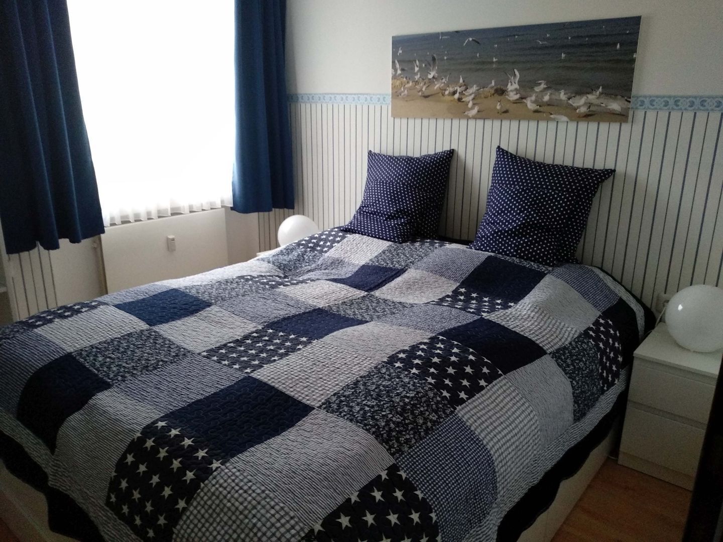 Ein Bett mit einer Decke mit Sternenmuster, zwei Kissen, ein Wandbild mit Vögeln und ein Nachttisch mit einem kugelförmigen Gegenstand.
