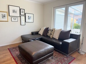 A living room with a dark leather sofa, a matching ottoman, and a patterned rug. There are framed pictures on the wall and a large window with blinds.