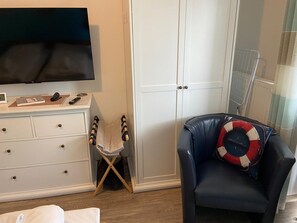 A hotel room with a TV, a desk with a chair, a wardrobe, a chair with a life preserver pillow, and a radiator.