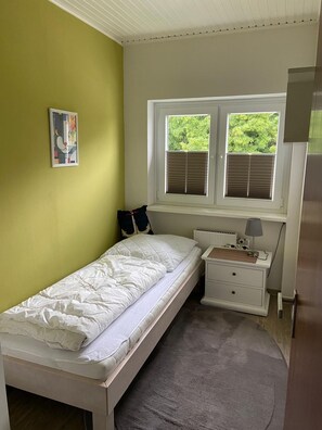 Quarto de casal com janela, radiador, mesa de cabeceira com luminária e travesseiro na cama.