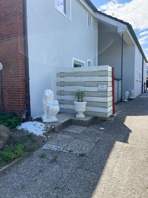 Edifício residencial com fachada branca, seção de tijolos à esquerda e cerca de madeira com uma placa. Há duas estátuas brancas, um vaso de plantas e um caminho de cascalho.