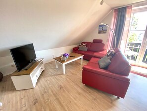 Un salon avec un téléviseur, une table basse en bois, un canapé rouge et une fenêtre avec des rideaux.