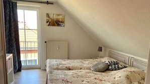 A bedroom with a bed, a radiator, a window with curtains, a painting on the wall, and a view of the outside. - 7016 "incl. beach chair" - 7016 "incl. beach chair" - An der Aue 9c - Apartment 25 (Dahme)