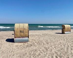 Two wicker beach chairs with metal bases on a sandy beach with the ocean in the background.