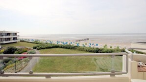 Blick von einem Balkon auf einen Strand mit einer Reihe blauer und weißer Liegestühle, einer Grünfläche und einem modernen Gebäude im Hintergrund.