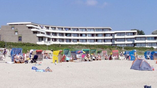 Ein Strand mit einem Hotel im Hintergrund, Liegestühlen und Sonnenschirmen.
