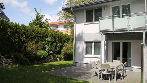 Une maison à deux étages avec un balcon, une terrasse avec une table et des chaises, et un mur de pierre agrémenté de verdure.